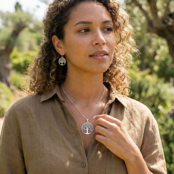 A woman with curly hair in a light brown shirt stands outdoors, touching her silver Tree of Life pendant necklace. She also wears matching Tree of Life earrings against blurred greenery and sunlight, evoking a peaceful, natural setting.