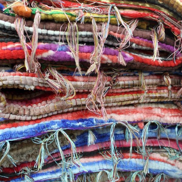 A close-up of the Large Velvet Rag Rug (105 × 185 cm), showing stacked, colorful woven stripes and patterns in red, blue, purple, brown, and cream with uneven, frayed threads—highlighting its handmade craftsmanship.