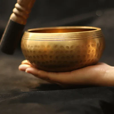 A hand holds the Nepal Tibetan Buddhist Singing Bowl Set | Handmade for Meditation, featuring a hammered texture and engraved lines. A wooden mallet hovers above, illuminated against a dark background to evoke calm and meditation.